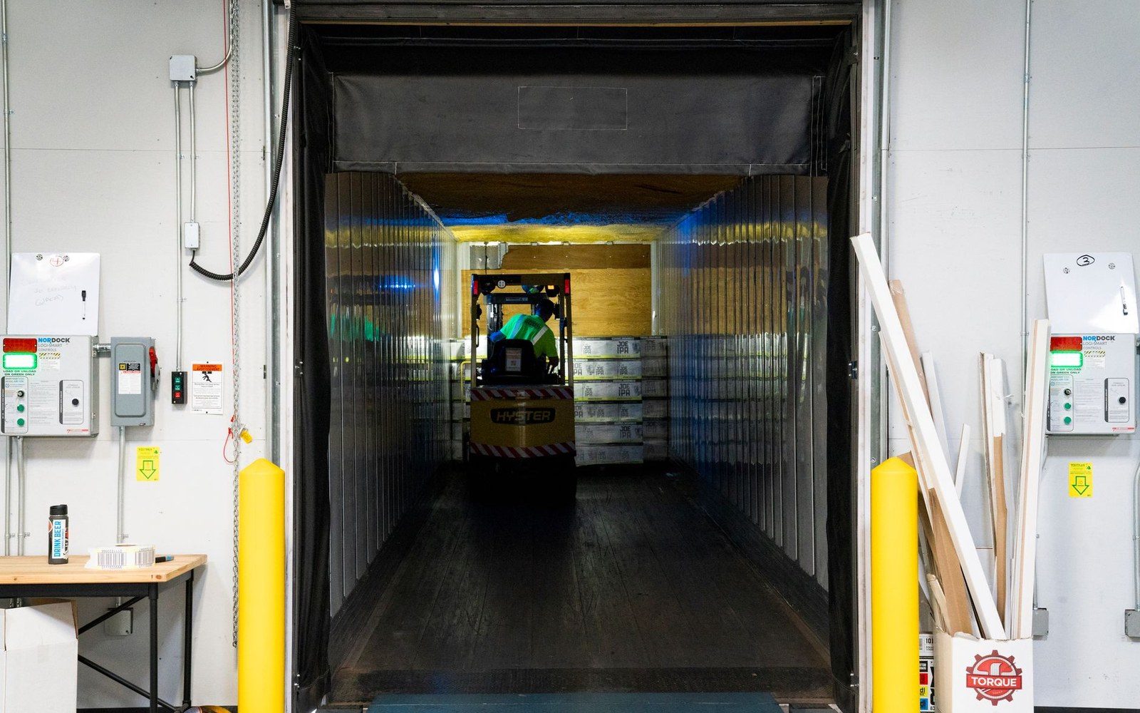 A forklift loads stacked boxes through plastic strip curtains into a truck bay