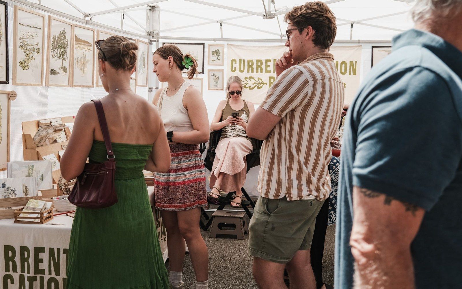 Visitors browse framed prints and cards at a crowded outdoor market tent booth