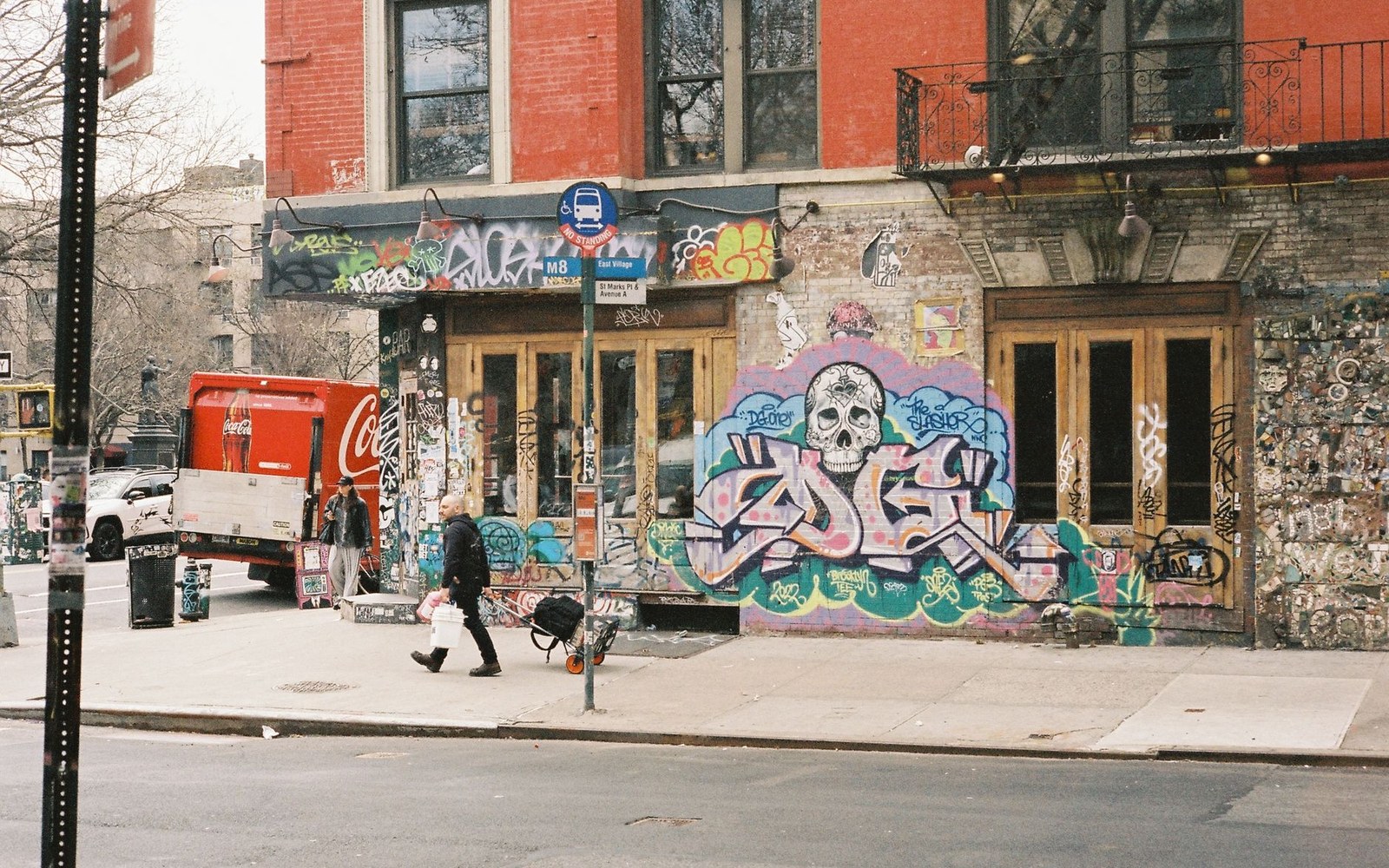 Graffiti-covered storefront with a skull mural as a pedestrian passes on the sidewalk