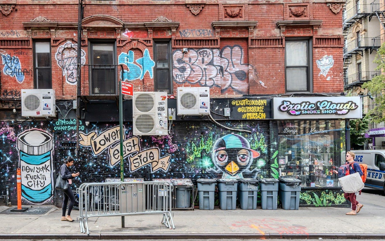 Graffiti-covered storefronts and trash bins line a Lower East Side street with pedestrians walking by